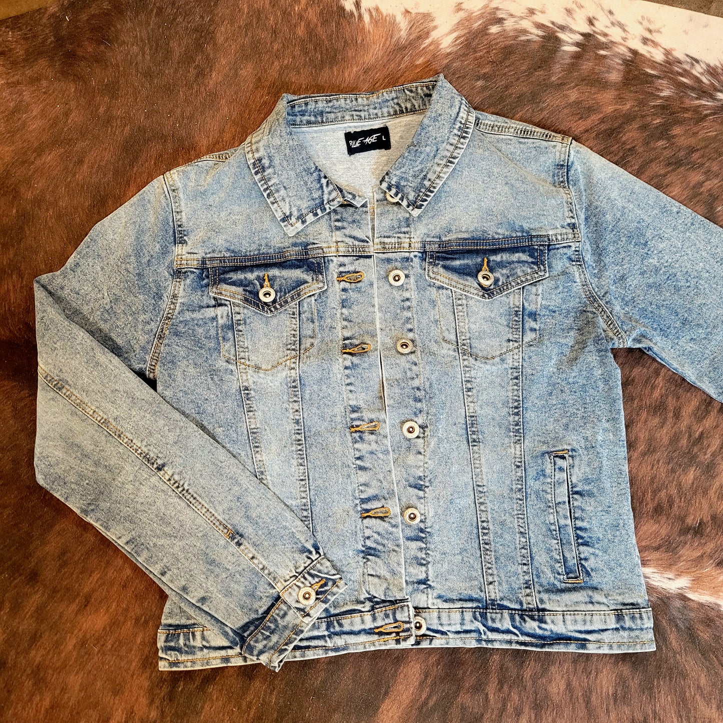 A light wash denim jacket with a button closure, displayed on a surface with a furry texture in the background.
