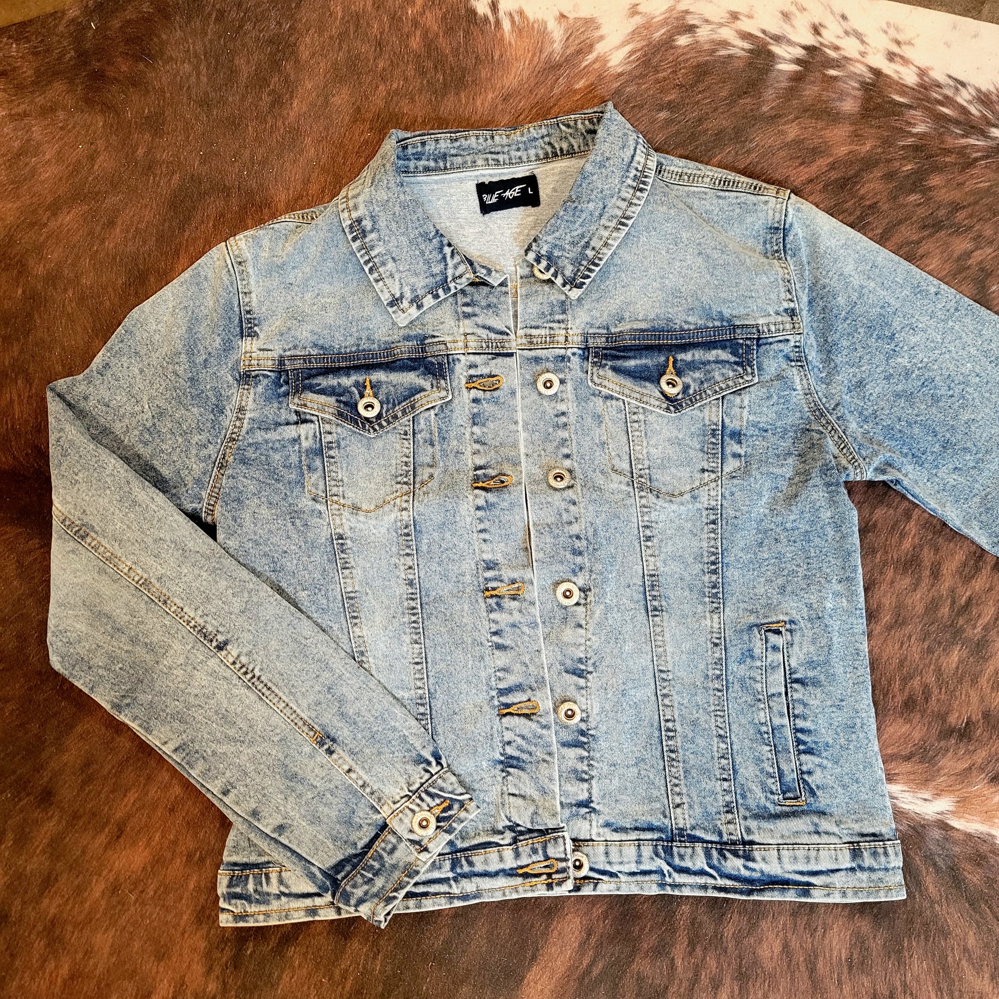 A light wash denim jacket with a button closure, displayed on a surface with a furry texture in the background.