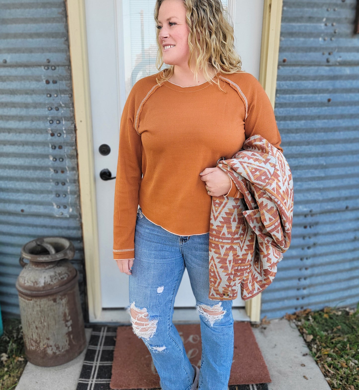 A woman wearing an orange waffle knit top with a round neckline, featuring exposed seams and contrast stitching. She has paired it with ripped jeans and is holding a patterned scarf.